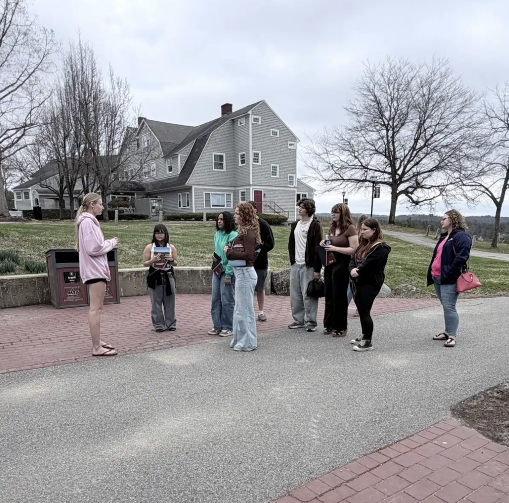 Students gather outdoors near a campus building as a guide speaks to them, with a large house-style building and open lawn in the background.