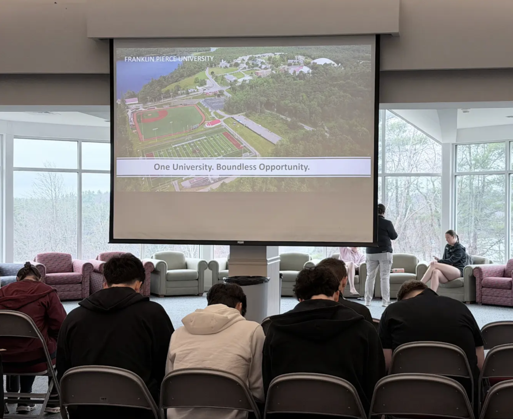 Students sit in rows of chairs indoors watching a presentation on a large screen displaying Franklin Pierce University campus information, with windows and lounge seating behind them.