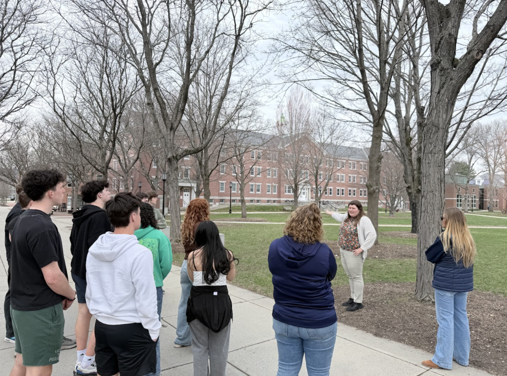 A group of high school students stands on a college campus walkway as a guide gestures toward buildings while speaking, with trees and a large brick academic building in the background.
