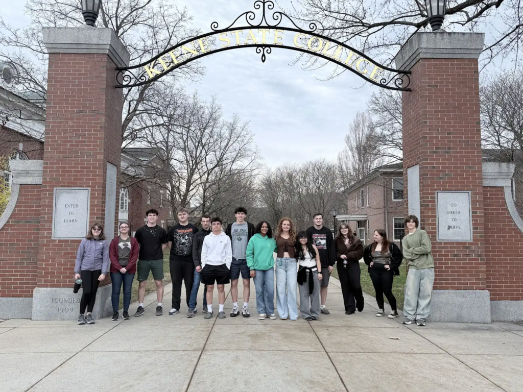 Students pose together under the Keene State College entrance arch, standing between two brick pillars on a campus walkway.