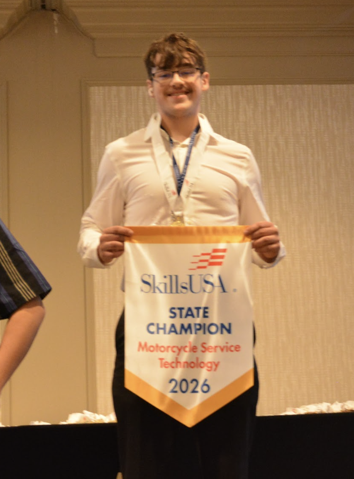 A student stands indoors wearing a medal and holding a SkillsUSA banner that reads “State Champion Motorcycle Service Technology 2026.”