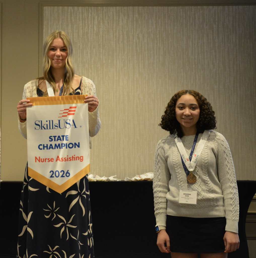 Two students stand side by side; one holds a SkillsUSA banner reading “State Champion Nurse Assisting 2026,” while the other wears a medal.