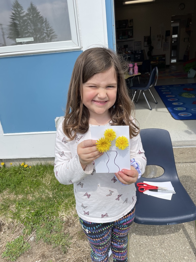 A young girl with long brown hair and a playful, scrunchy-faced expression holds her dandelion art close to the camera. Her card features a wavy-lined vase with three dandelions. She is wearing a white long-sleeved shirt with butterfly patterns and colorful patterned leggings.  