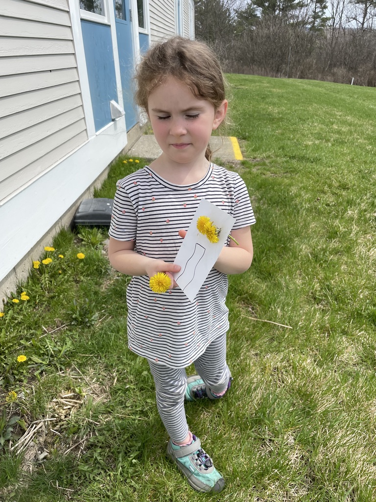  A young girl with reddish-brown hair in a striped shirt stands in a grassy area next to a building. She is looking down thoughtfully at a small white card she is holding, which features a hand-drawn vase with two yellow dandelions poked through it to look like a bouquet.