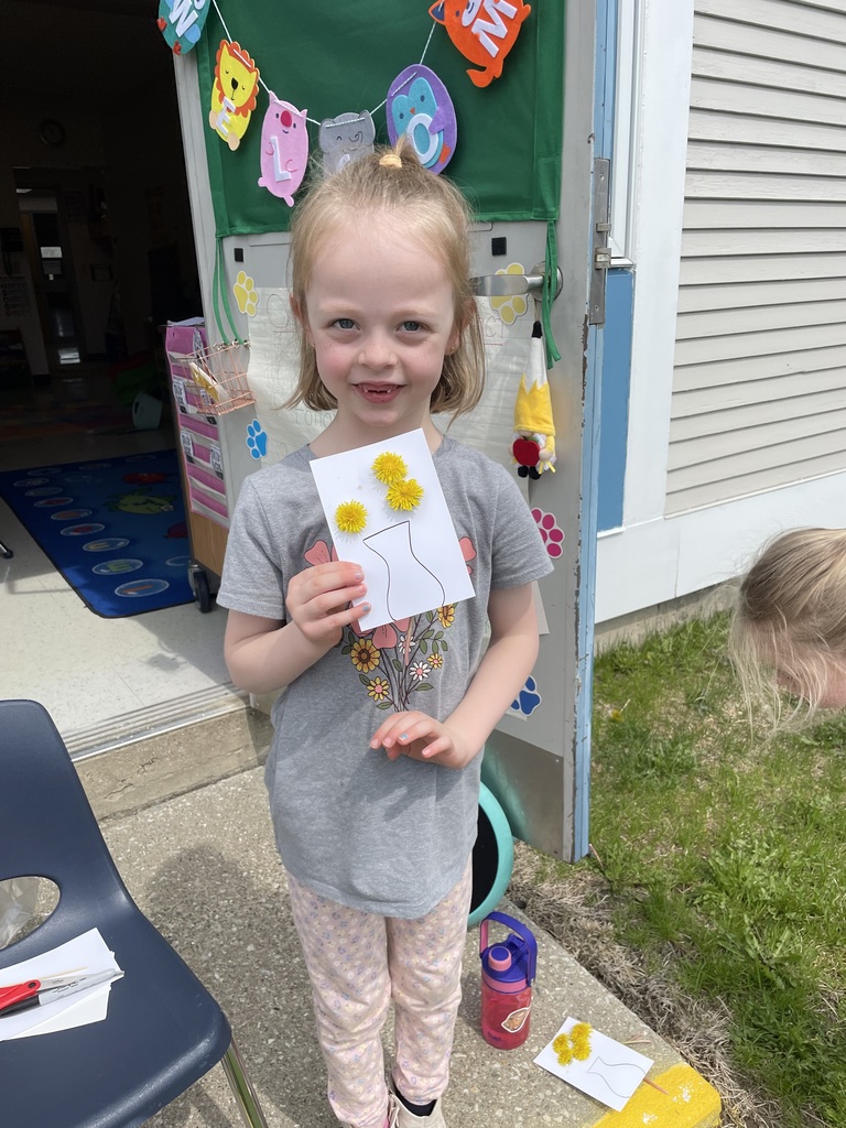 : A smiling girl with blonde hair tied in a small top-knot stands in a doorway, proudly holding up her artwork. Her white card shows a drawing of a vase holding three bright yellow dandelions. She is wearing a grey t-shirt with a floral graphic and patterned pink leggings.  