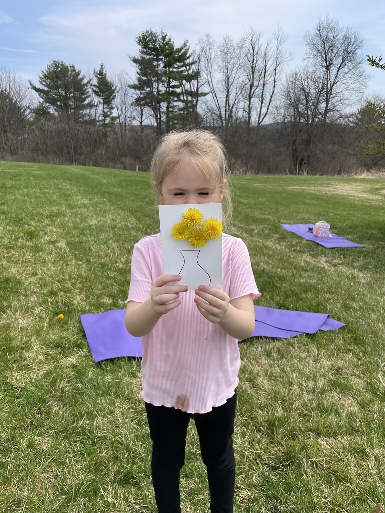 A young girl in a pink ribbed shirt and black leggings stands in a wide green field, holding her dandelion craft directly in front of her face. The white card shows four yellow dandelions "planted" in a hand-drawn vase. In the background, purple yoga mats are spread out on the grass.  