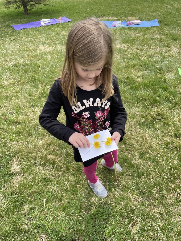  A high-angle shot of a young girl with light brown hair wearing a black "Always" butterfly shirt and bright pink leggings. She is carefully adjusting yellow dandelions on a white card with a vase drawing while standing in a grassy field. Yoga mats and water bottles are visible in the background.