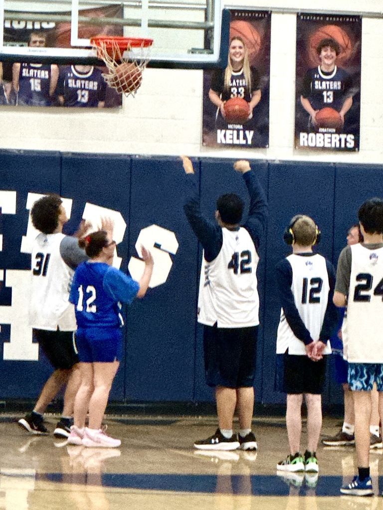 Several players stand under the basket watching the ball approach the hoop during a game, with banners and posters visible on the wall behind them.