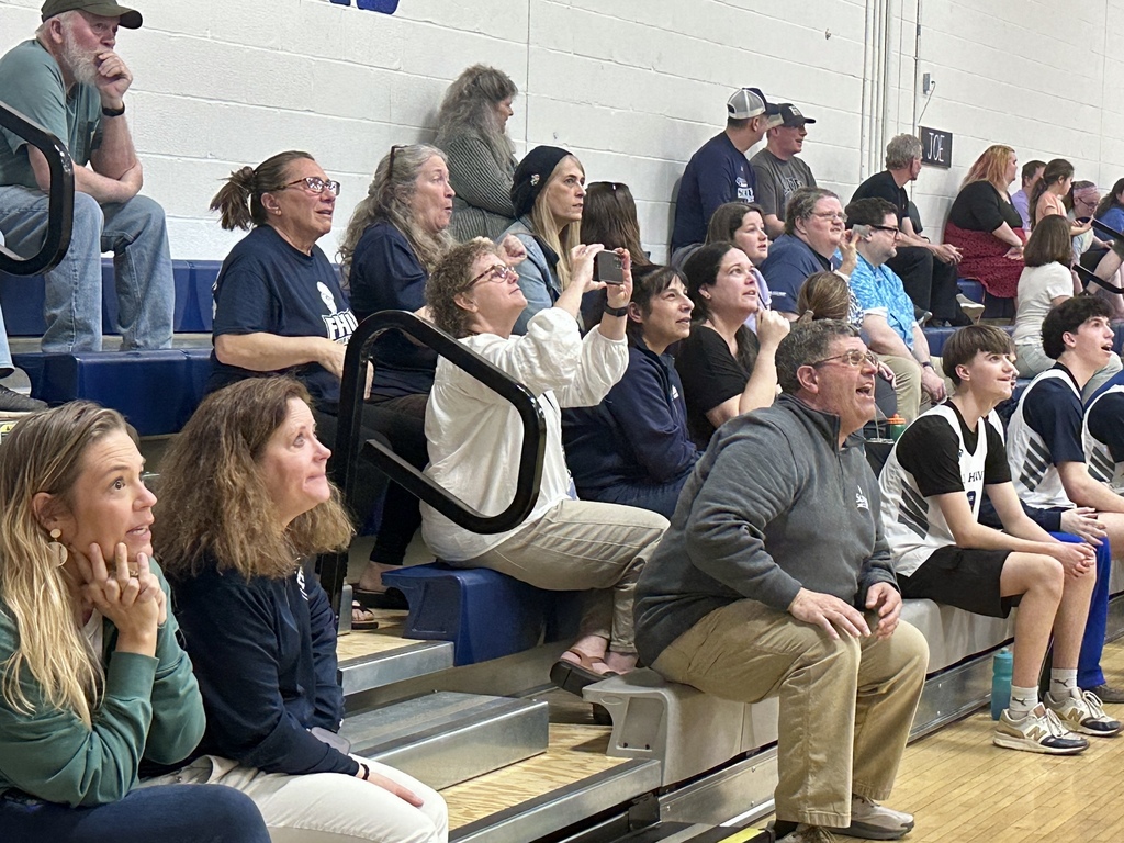 A crowd of spectators sits on bleachers, watching the game intently; some are smiling and taking photos.