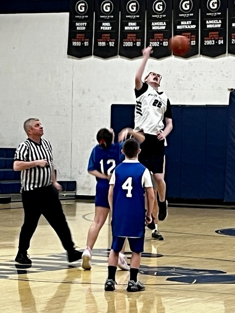 A player in a white jersey jumps to shoot the basketball while others look on, with a referee nearby during the game.
