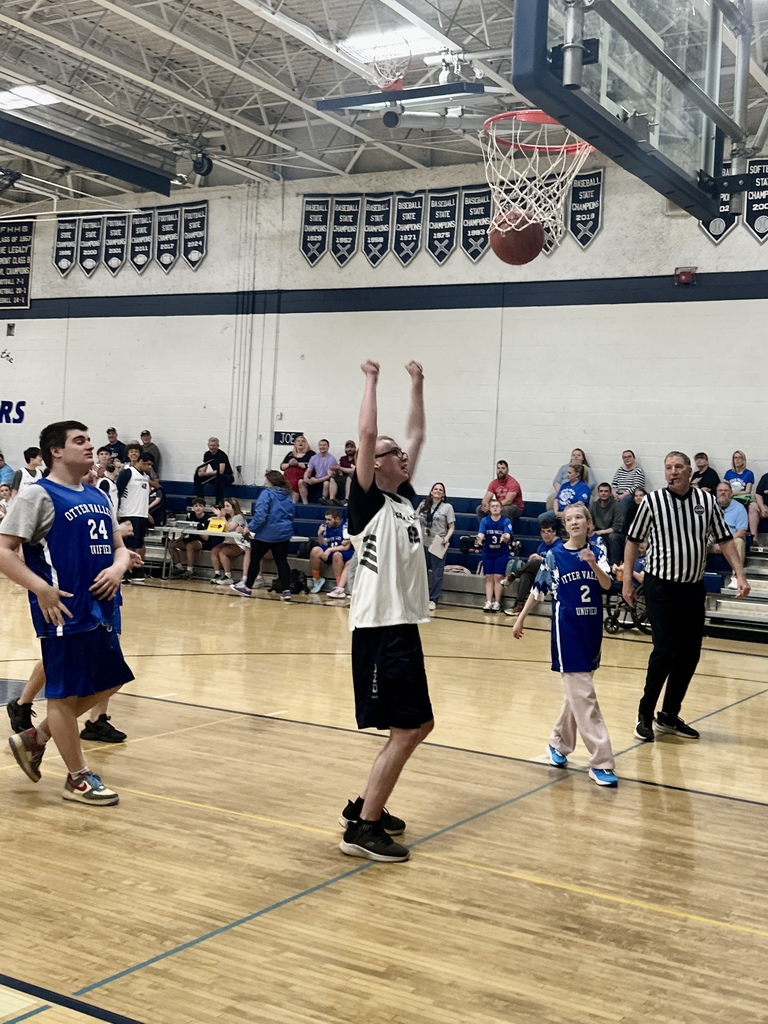 Student in a white jersey celebrates with arms raised as a basketball goes through the hoop during a Unified Basketball game, with players and spectators in the background.
