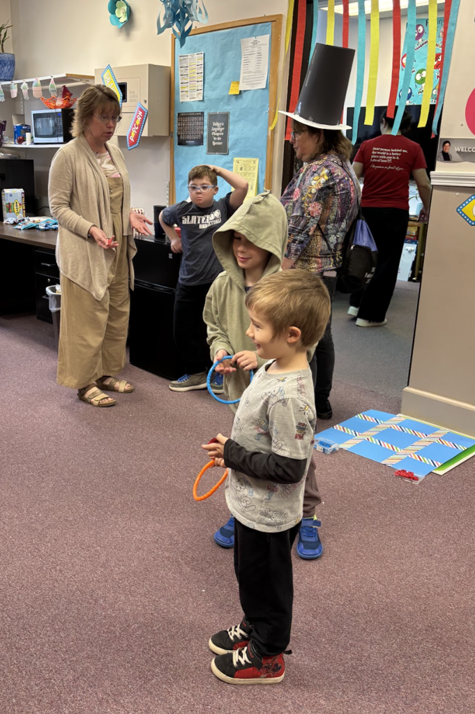Students participate in a classroom carnival game, holding colorful rings while a teacher and another adult supervise; decorations and streamers hang in the background.