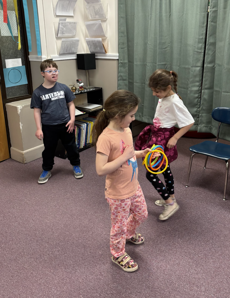 Three students play a ring toss-style game indoors, with one student holding several colorful rings while others take turns participating.