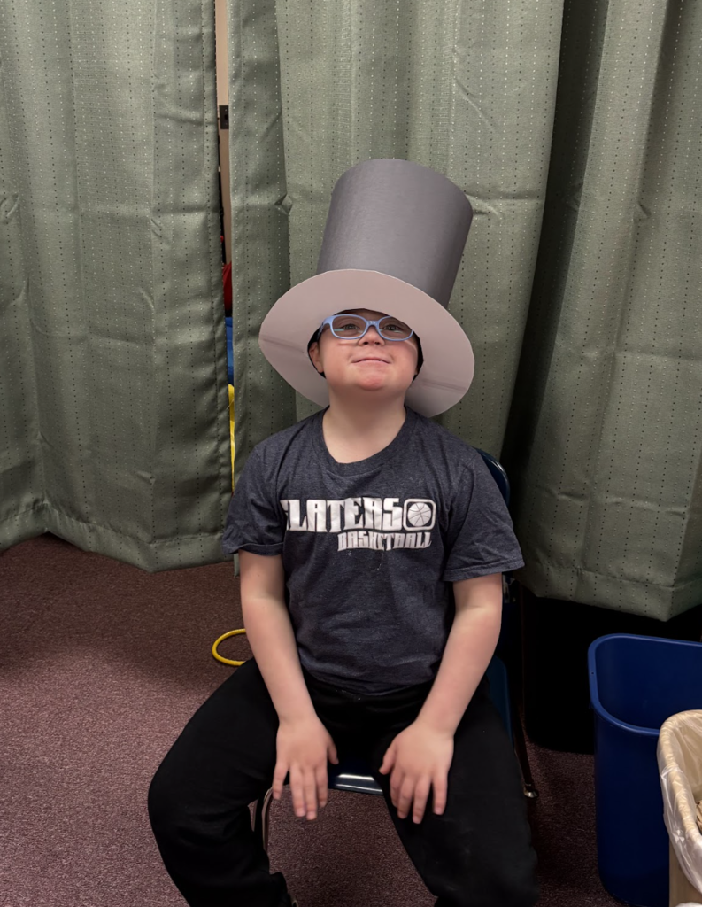 A student wearing an oversized top hat sits on a chair, smiling during the classroom carnival activity.