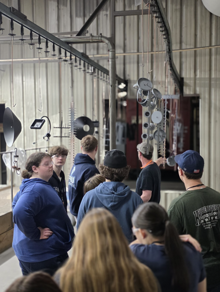 Students walk through a manufacturing area while observing hanging metal parts moving along a production line.