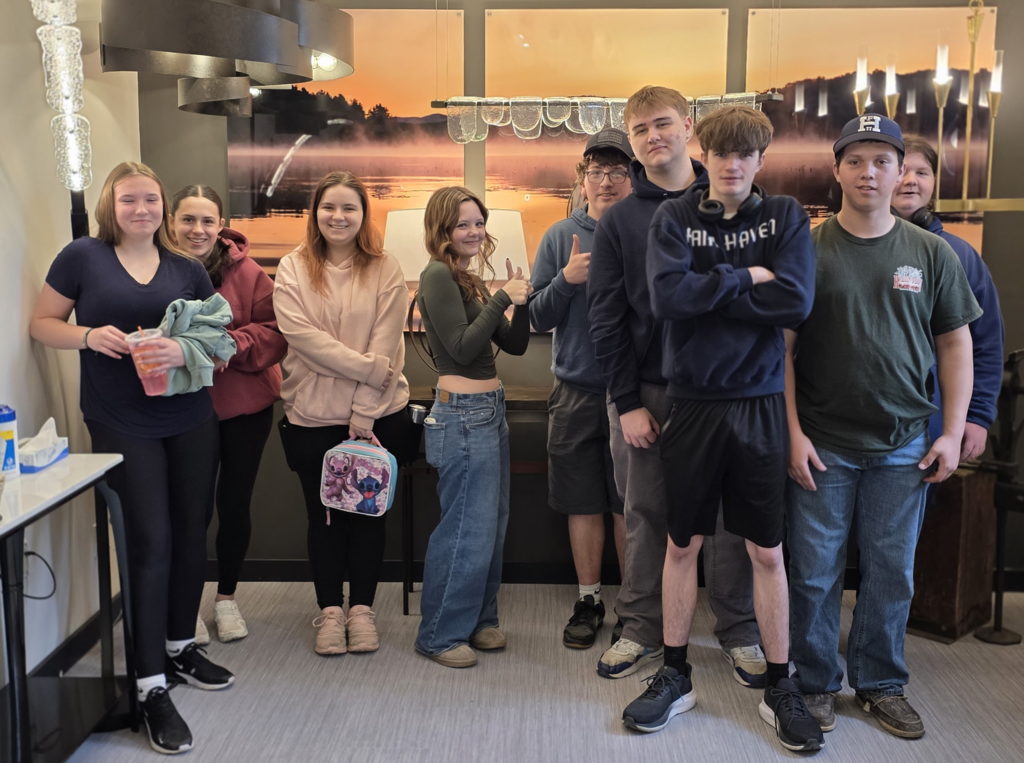 A group of high school students pose together indoors, smiling in front of a display featuring decorative lighting fixtures.