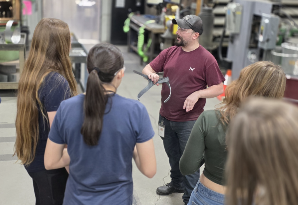 An employee holds a metal component while speaking to students gathered around in a workshop.