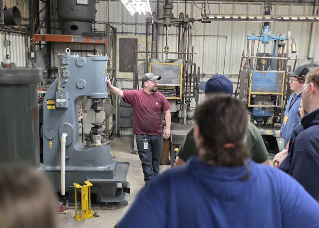 An employee stands beside large industrial machinery, explaining equipment and processes to a group of students.