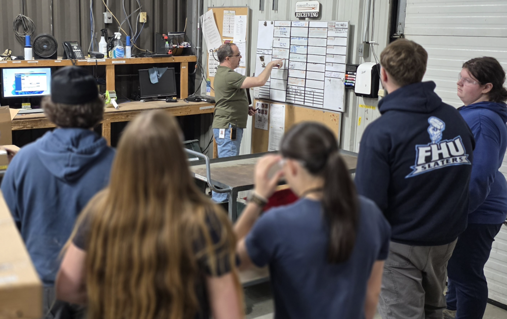 Students stand in a workshop listening as an employee points to a scheduling board and explains production processes.