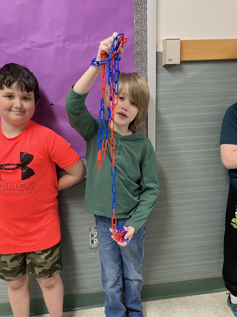  A medium shot of a young boy with blonde hair holding up a tangled bunch of red and blue plastic chains. He is wearing a green long-sleeved shirt and jeans. To his left, another boy in a red Under Armour shirt smiles at the camera while standing against a purple bulletin board. 