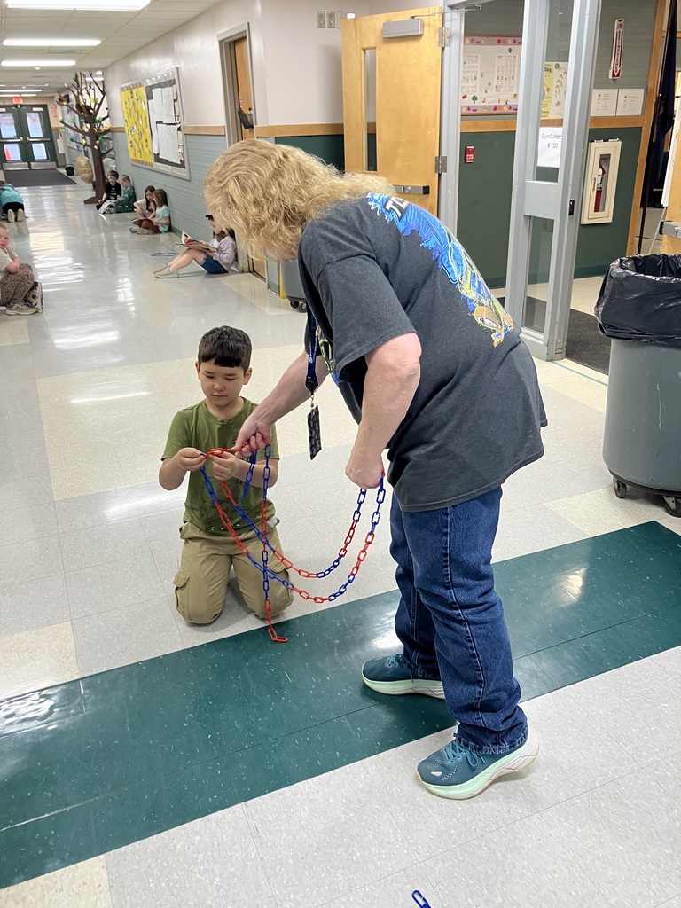 ) In a school hallway, an adult with curly blonde hair leans over to help a young boy in a tan outfit connect sections of a red and blue plastic chain. Other children are visible sitting along the walls in the background. 