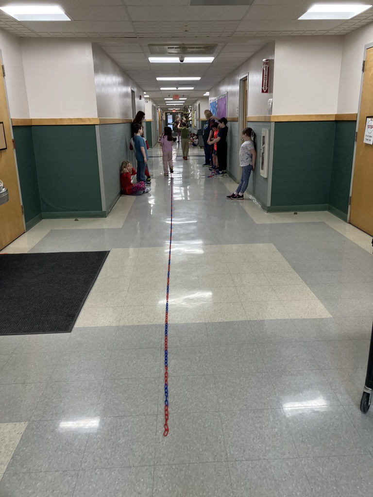 A wide, long-distance shot down a bright school hallway. A very long chain of red and blue plastic links stretches down the center of the floor, extending toward the far end of the hall. Several students and staff members are scattered along the sides of the hallway, watching or participating in the activity.