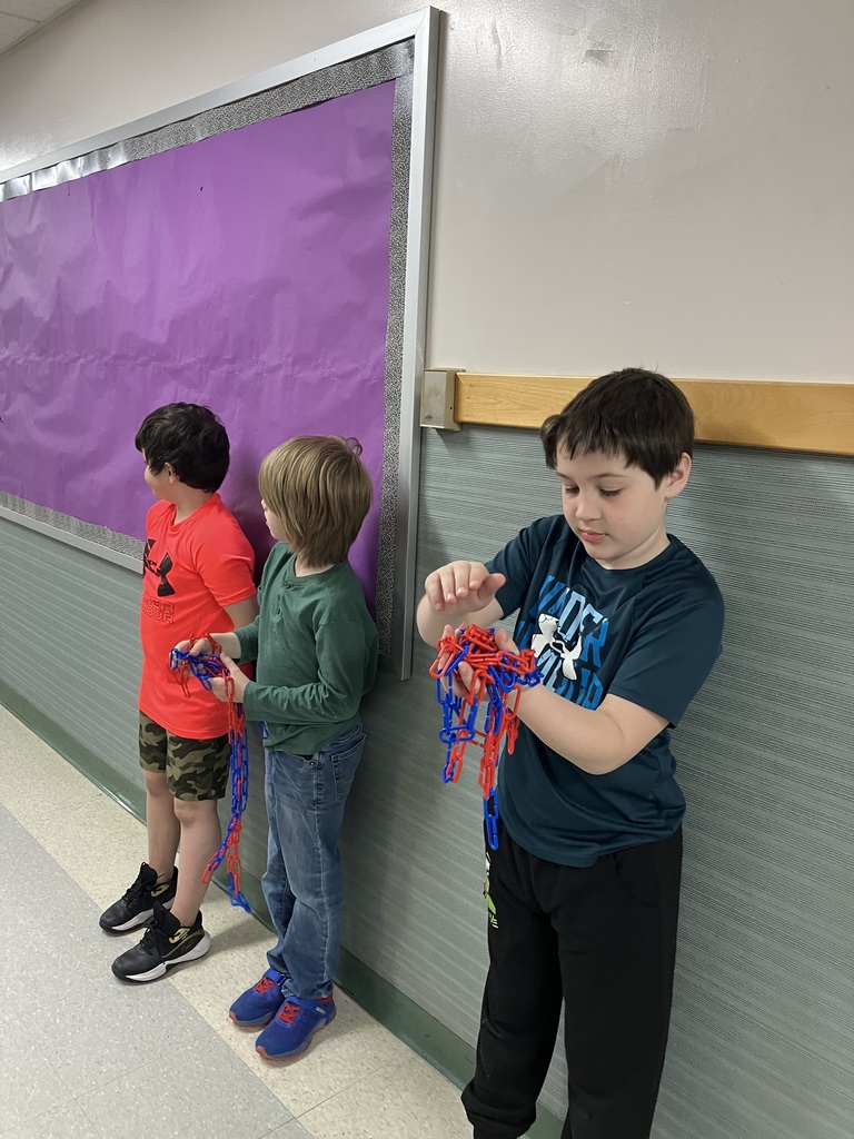  Three young boys stand against a hallway wall next to a purple bulletin board, each holding or playing with lengths of red and blue plastic chains. The boy in the foreground, wearing a dark teal shirt, is focused on gathering the chain in his hands.  