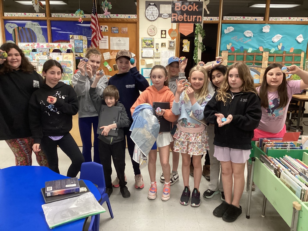 Larger group of students stand together in a classroom, proudly displaying their colorful painted rocks.