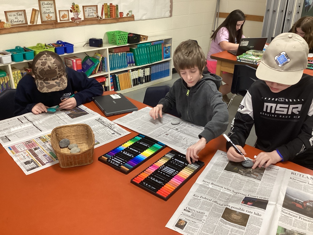 Three students sit at a classroom table painting rocks, with colorful markers, newspapers, and a basket of stones in front of them.