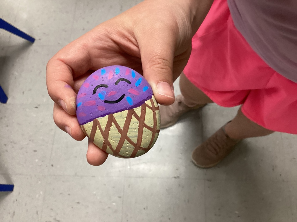 Close-up of a hand holding a painted rock with a smiling purple top and decorative patterns in pink and blue.