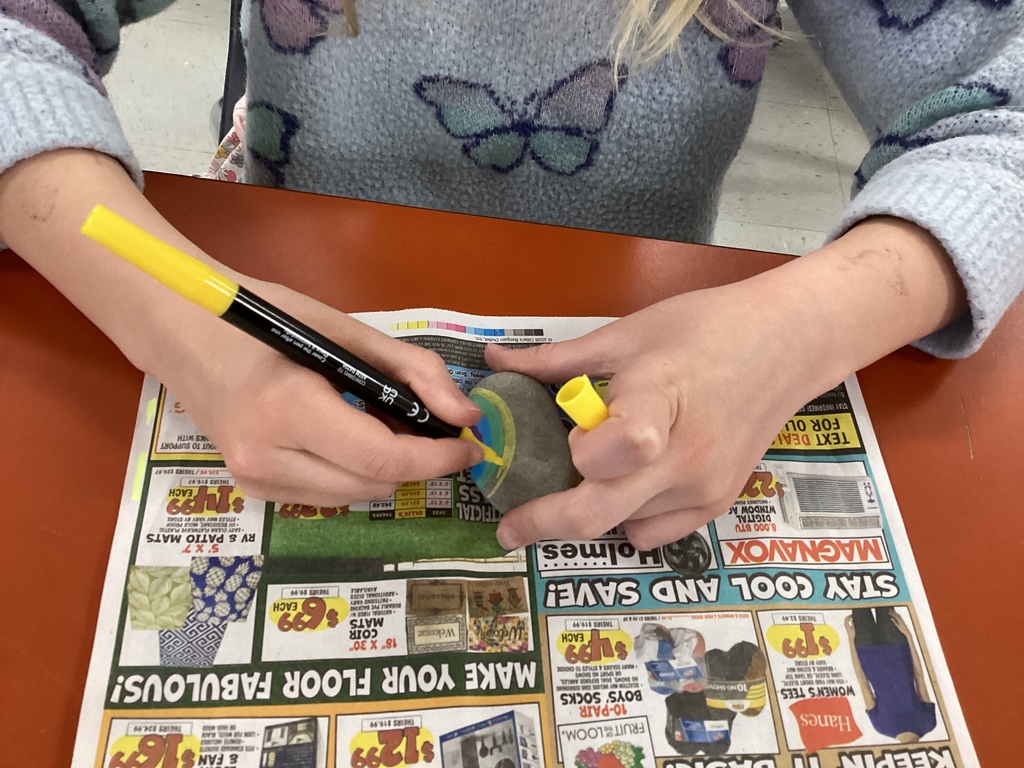 Student’s hands use a yellow paint marker to create a colorful swirl design on a smooth gray rock, with newspaper protecting the table underneath.