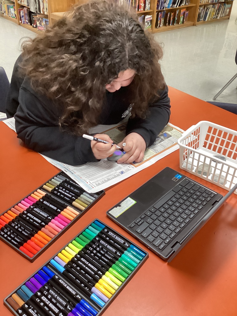Student leans over a table painting a rock with a white marker; trays of bright paint markers and a closed laptop sit nearby.