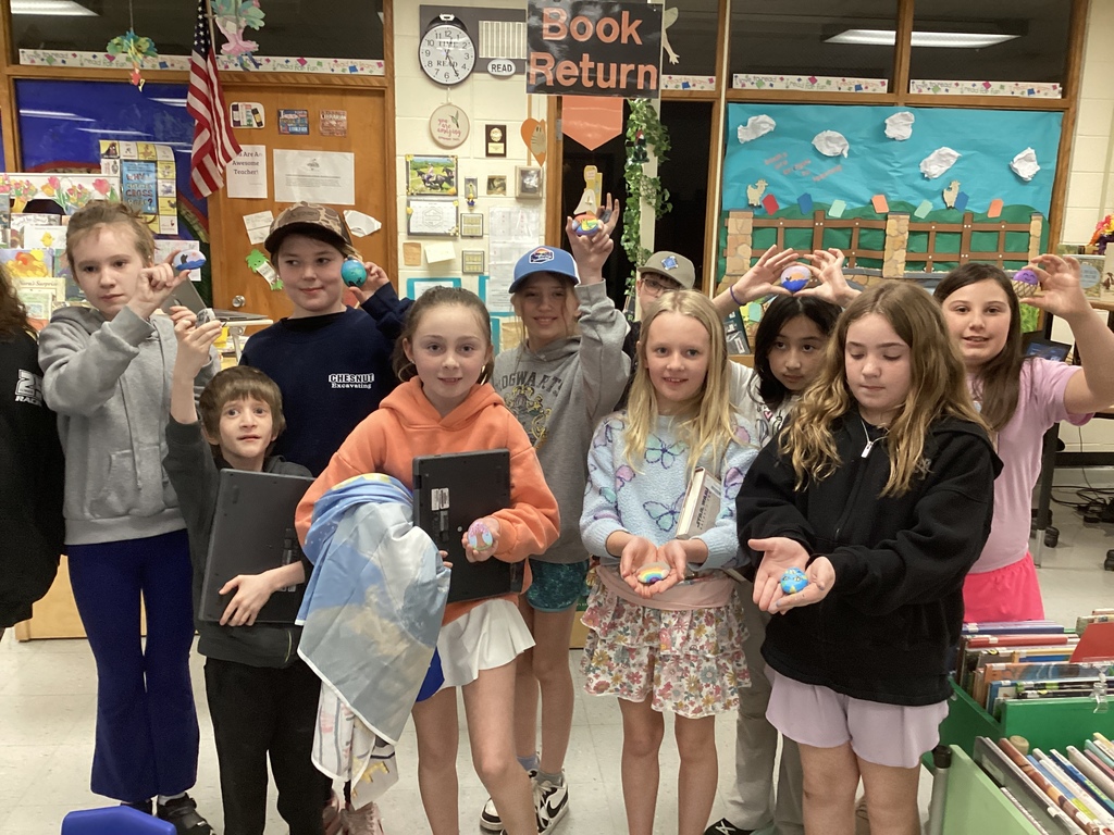 Group of elementary students in a classroom hold up their painted rocks, smiling and showing their designs.