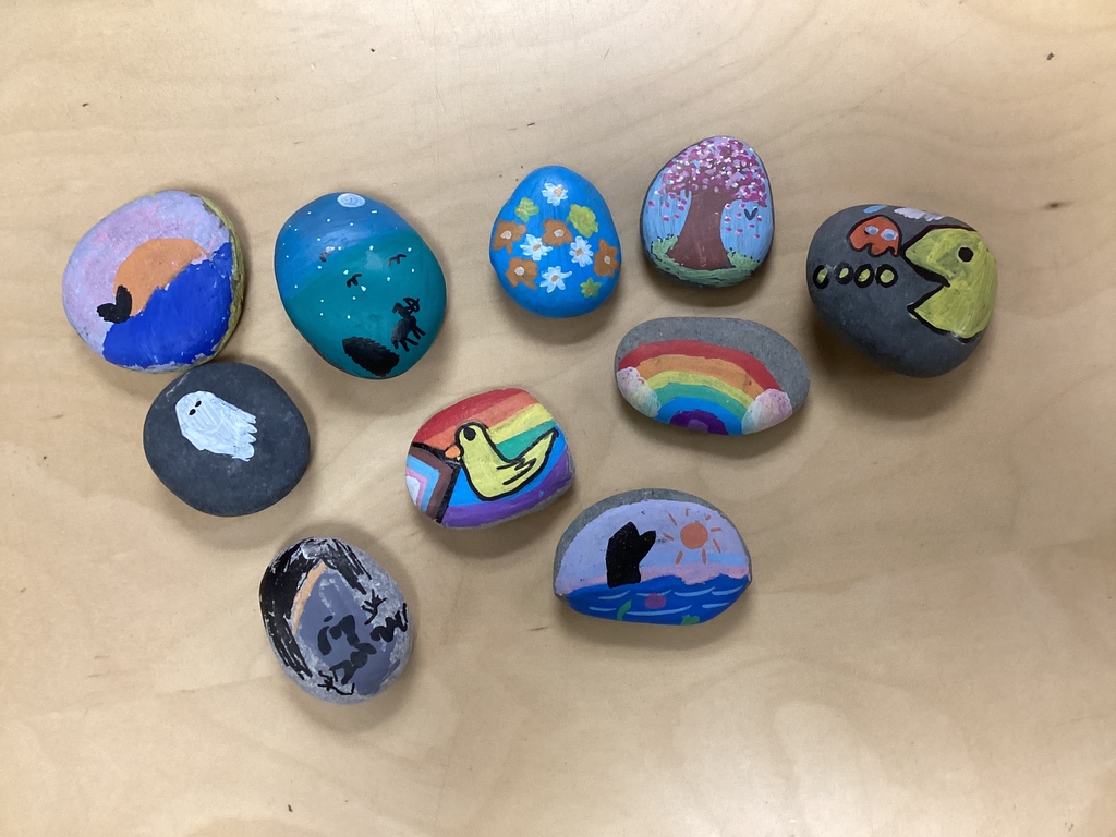 A group of decorated rocks displayed on a table, featuring designs such as rainbows, flowers, a tree, a bird, and a sunset scene.