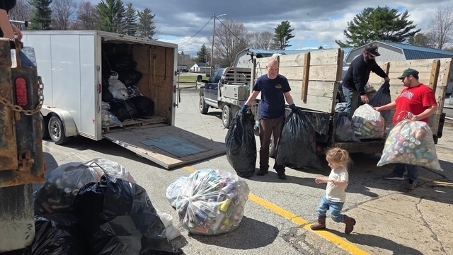 Volunteers carry heavy bags of bottles and cans toward a trailer while a small child walks nearby in the parking lot.