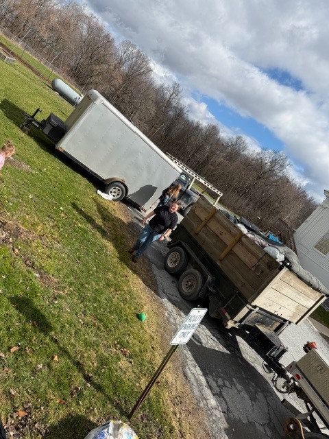 Truck and trailer filled with bags of recyclables parked near a grassy area, with people standing nearby under a partly cloudy sky.