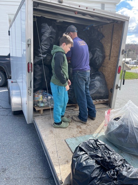 Adult and child stand inside an open trailer stacking bags of recyclables, with more bags piled around them.