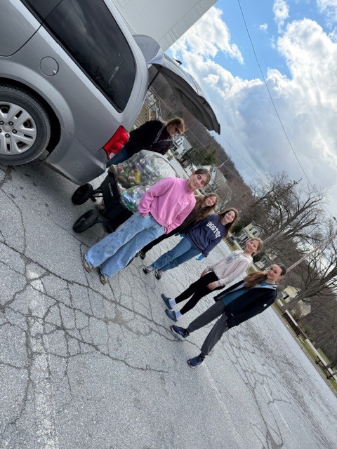 Group of students stand next to a vehicle holding bags of collected bottles and cans, smiling in a parking lot.