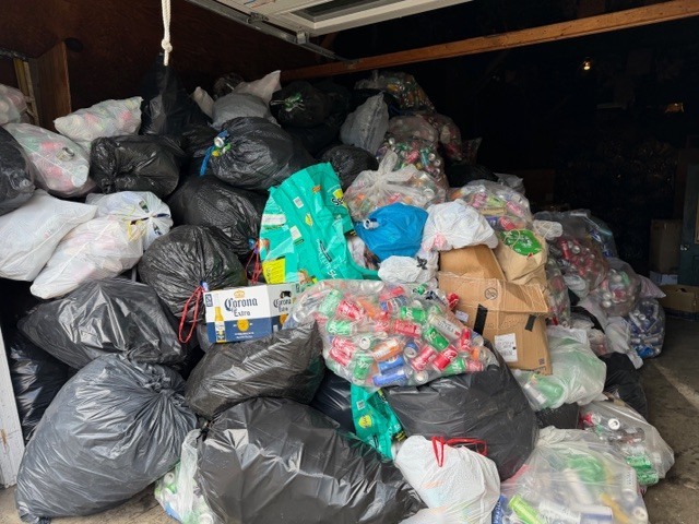 Large pile of black trash bags and clear bags filled with cans and bottles stacked high inside a storage space.