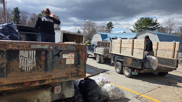 People load large bags of recyclables into a truck and trailer in a parking lot, with piles of filled bags on the ground nearby.