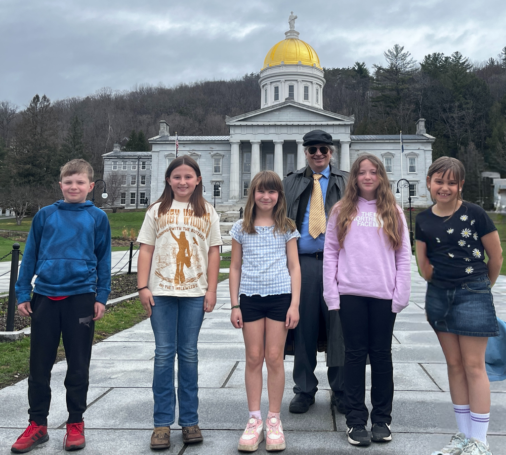 Group of five elementary students and an adult stand in front of the Vermont State House with its gold dome, posing on a walkway with trees and hills in the background.