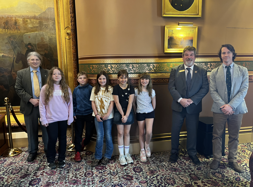 Group of students stand indoors with two adults in suits inside the Vermont State House, posing in front of framed artwork and decorative walls.