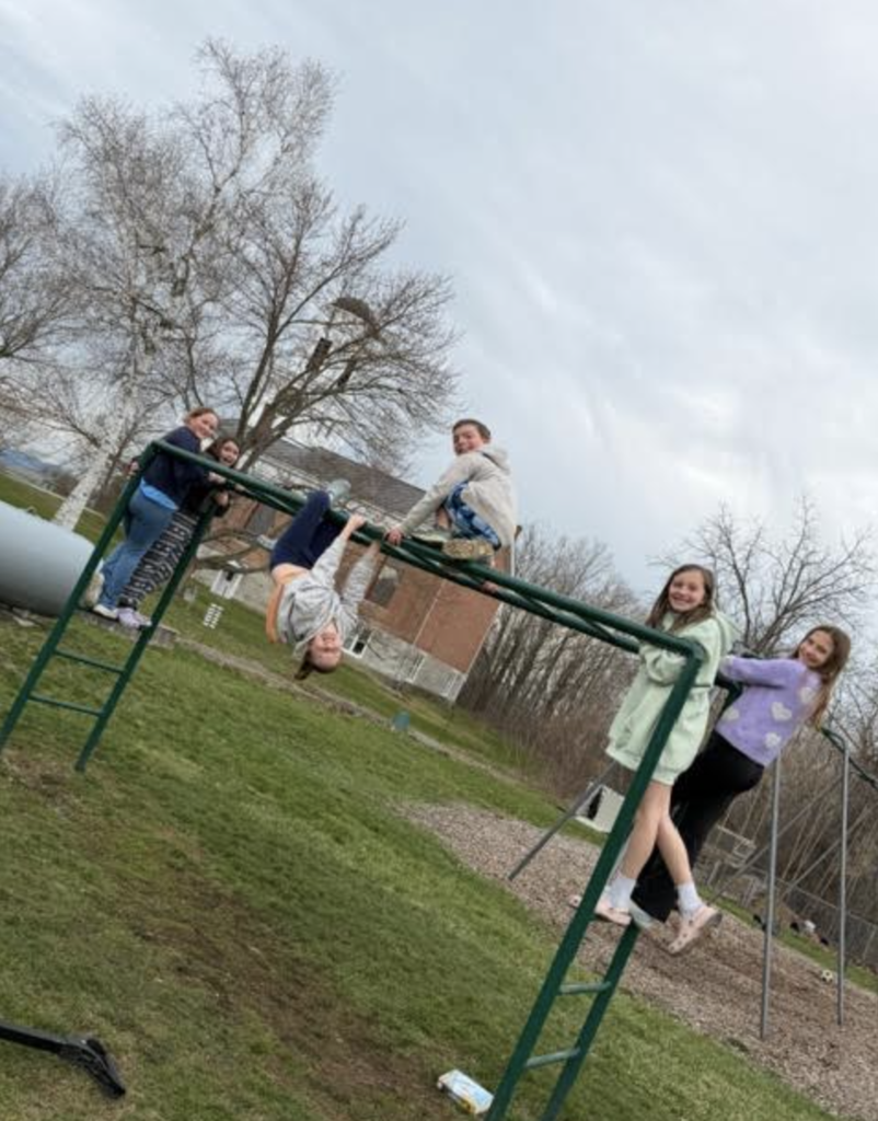 Students climb and hang from monkey bars, with one student upside down; playground equipment and trees are visible nearby.