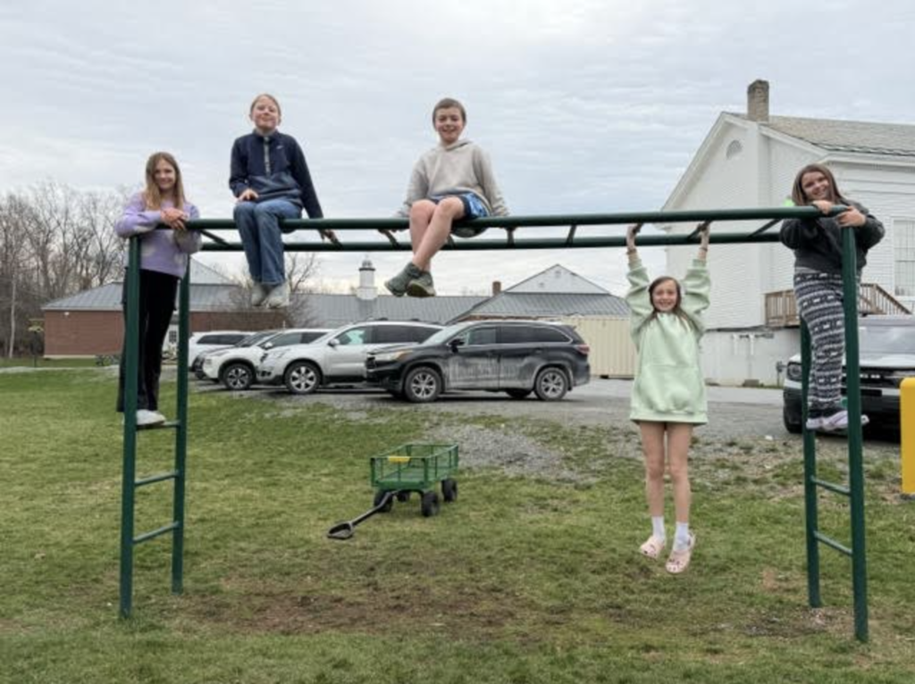 Five students play on monkey bars outside, with some sitting on top and one hanging below; parked cars and a building are visible in the background.