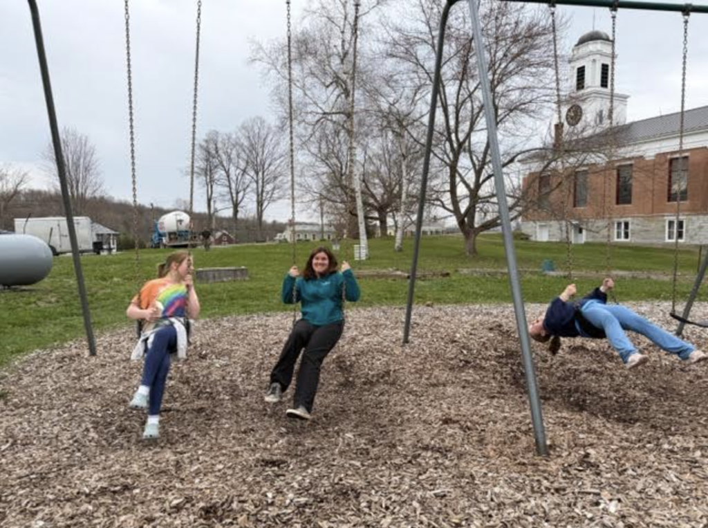 Three students swing on a playground set while another leans back on a swing; a school building and leafless trees are visible behind them.