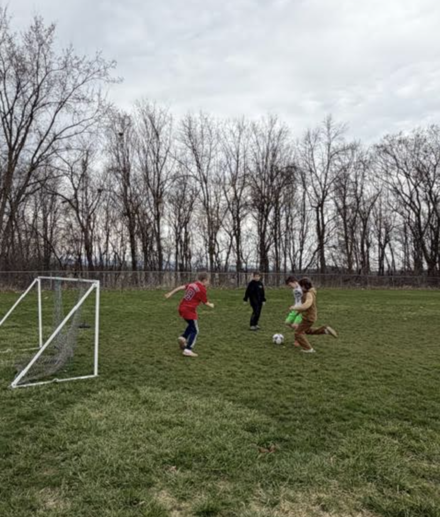Four students play soccer on a grassy field near a small goal, with trees lining the background.