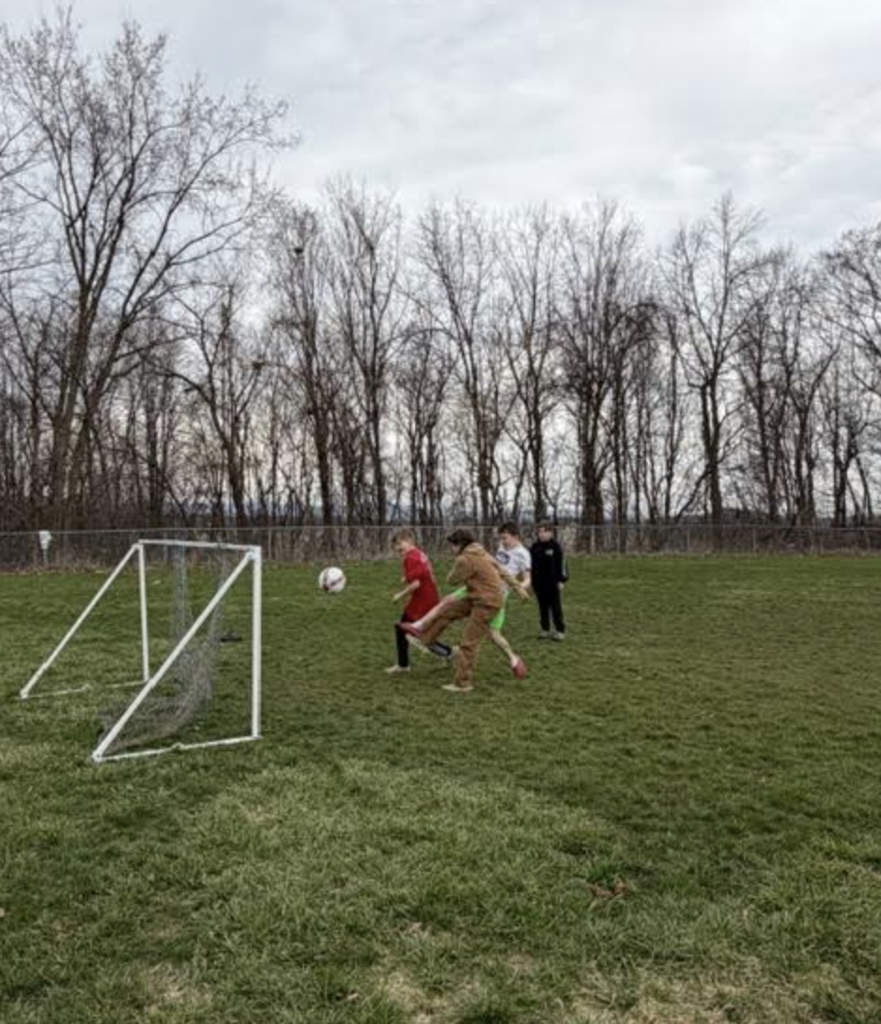 Students kick a soccer ball toward a small goal during a game on the grass field, with trees in the background.