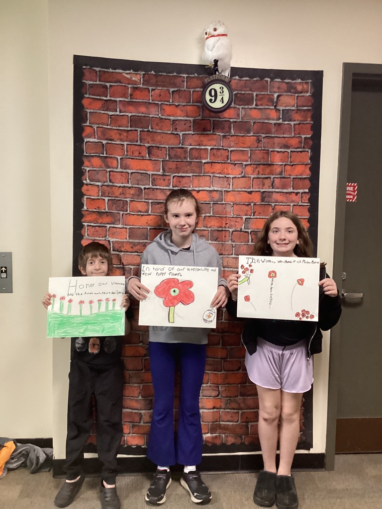 Three students stand in front of a brick wall display holding hand-drawn poppy posters honoring veterans.