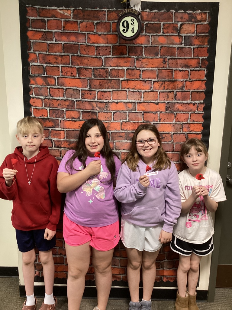 Four students stand together holding small red poppies, smiling in front of a brick wall display.