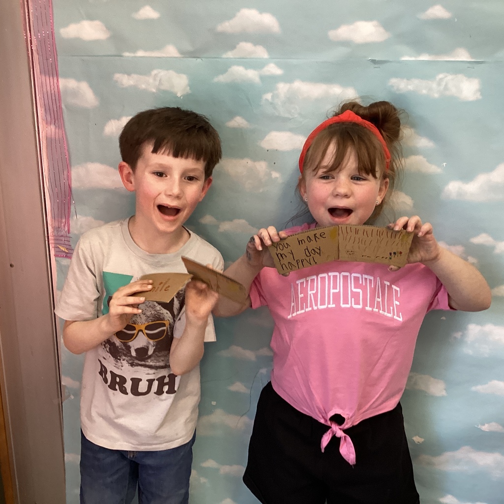 Two students smile and hold up handmade coffee sleeve cards with cheerful messages, including “You make my day happy,” against a sky-and-cloud background.